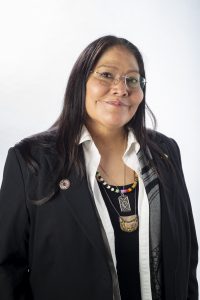 A woman with long dark hair and glasses wears a black blazer, white shirt, and several beaded necklaces. *AI generated alt text