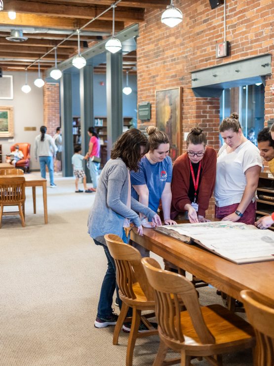 Six people stand around a long wooden table in a museum, looking down at a large open book. *AI generated alt text