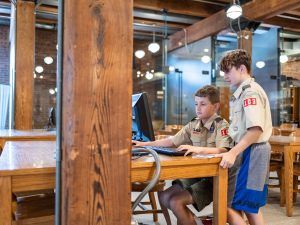Two scouts in uniform stand at a wooden desk in a museum, one typing on a computer while the other watches the screen. *AI generated alt text