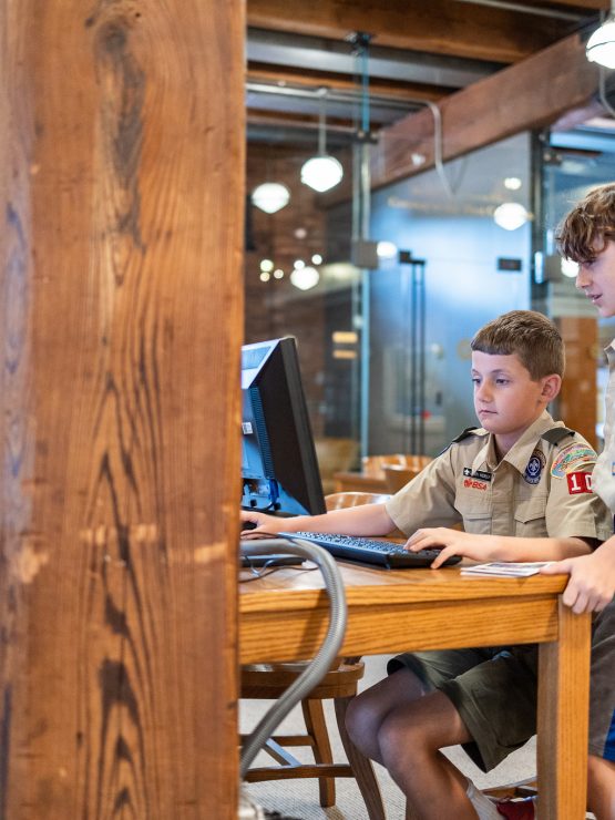 Two scouts in uniform stand at a wooden desk in a museum, one typing on a computer while the other watches the screen. *AI generated alt text