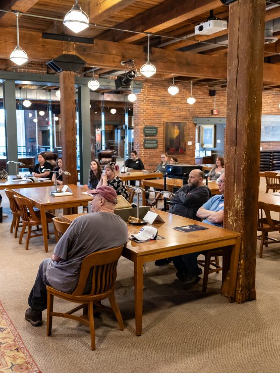A woman in a red dress speaks to a group of people seated at wooden tables in a museum hall featuring exposed ceiling beams and brick walls. *AI generated alt text