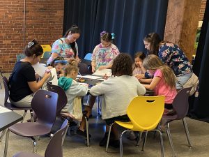 A scout troop of adults and children sit around a table at the museum crafting with paper and glue. *AI generated alt text