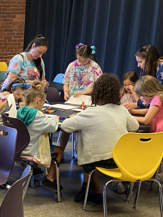 A scout troop of adults and children sit around a table at the museum crafting with paper and glue. *AI generated alt text