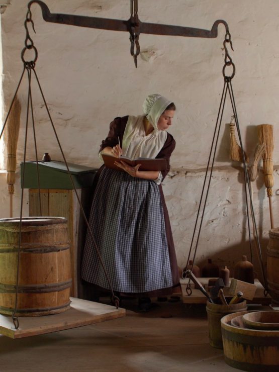 A woman in historical colonial clothing writes in a ledger at a museum exhibit while standing behind a large wooden hanging scale. *AI generated alt text