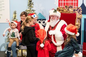 A group of children in festive clothes gather around Santa at the museum, where Santa is seated in a large red chair. An adult stands nearby, smiling, with decorated trees and holiday decorations visible in the background. *AI generated alt text