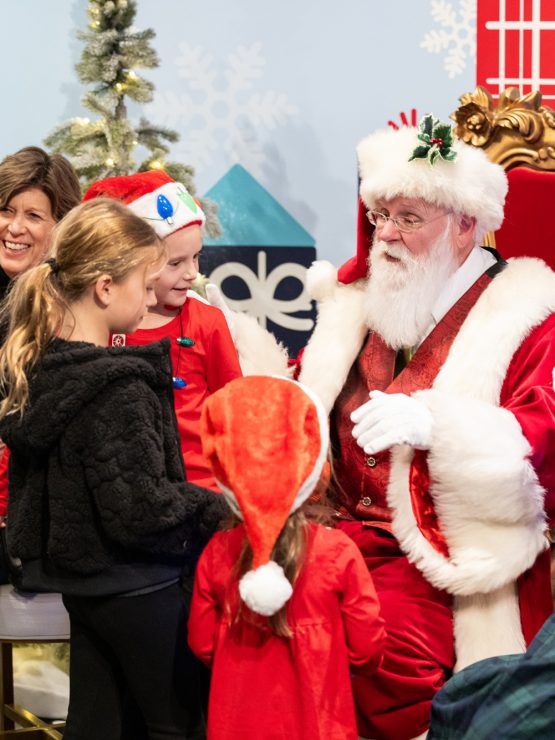A group of children in festive clothes gather around Santa at the museum, where Santa is seated in a large red chair. An adult stands nearby, smiling, with decorated trees and holiday decorations visible in the background. *AI generated alt text